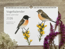 Lade das Bild in den Galerie-Viewer, Vogelkalender 2026, auf dem Deckblatt Schwarzkehlchen auf gelben Blüten. Als Deko liegen einige Blumen neben dem Kalender